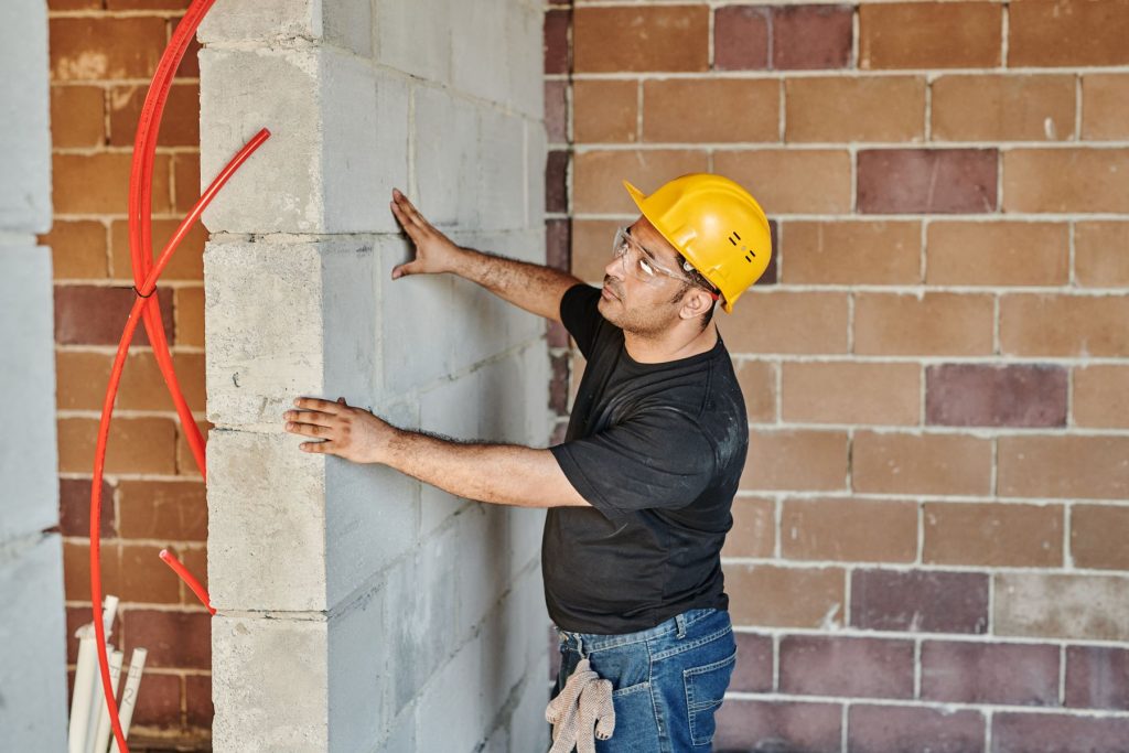 Bauarbeiter mit Schutzhelm prüft eine Rohbauwand mit verlegter Heizungs- und Wasserleitung