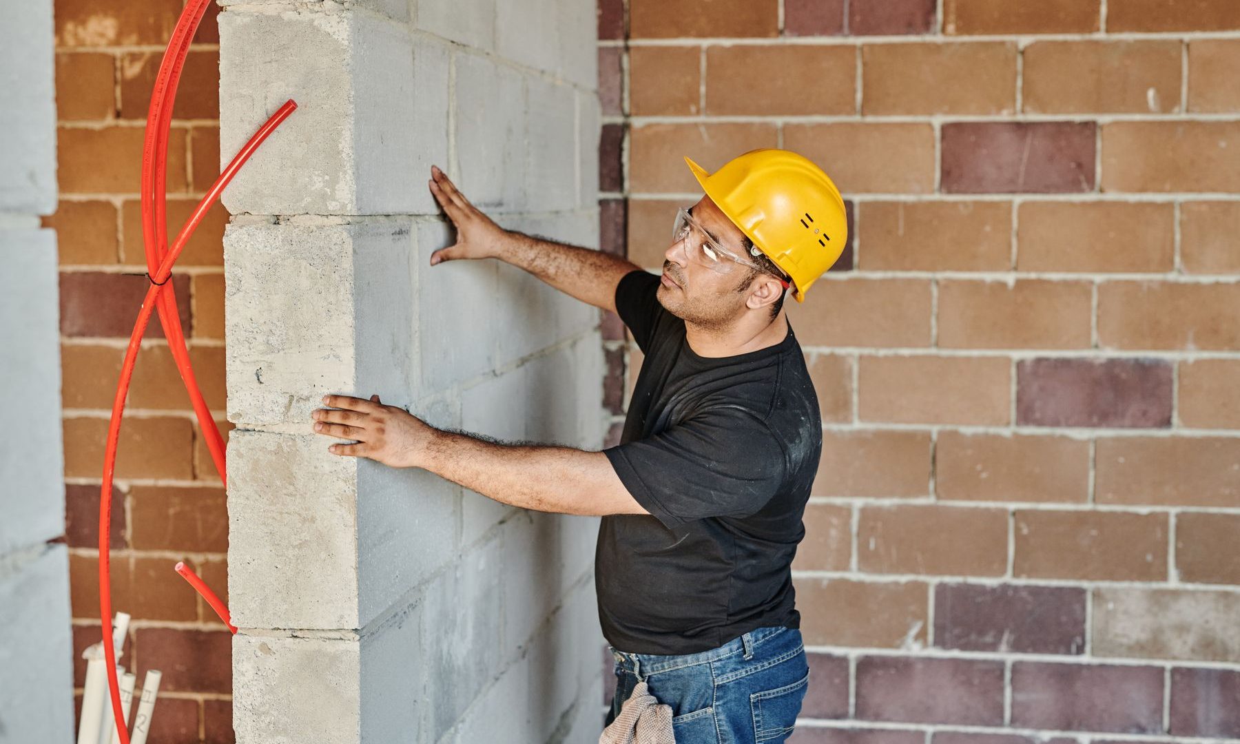 Bauarbeiter mit Schutzhelm prüft eine Rohbauwand mit verlegter Heizungs- und Wasserleitung